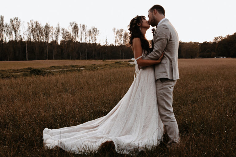 Zu sehen ist ein Brautpaar bei einem Brautpaarshooting in Garmisch-Partenkrichen, sie stehen auf einem Feld im Sonnenuntergang und küssen sich. Das Kleid der Braut ist über dem Boden ausgebreitet und man sieht die Liebe zwischen den beiden, aufgenommen in der Dämmerung in Farbe