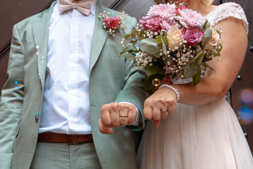 Ein Brautpaar hält nach der Trauung die Fäuste in die Kamera, man sieht die Eheringe. Das Bild ist bunt und man sieht den Blumenstrauß