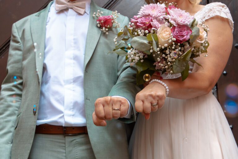 Ein Brautpaar hält nach der Trauung die Fäuste in die Kamera, man sieht die Eheringe. Das Bild ist bunt und man sieht den Blumenstrauß