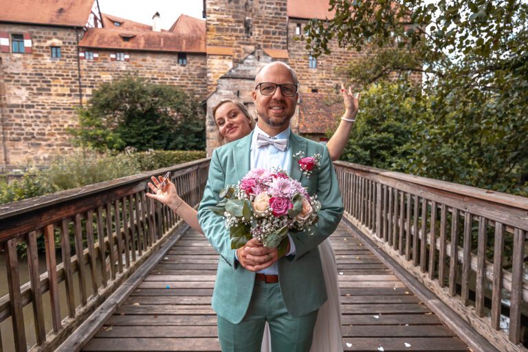 Ein Brautpaar steht auf einer Brücke in Bayern und sie lachen in die Kamera, sie sind sehr glücklich. Die Braut steht hinter dem Bräutigam und schaut ihm über die Schulter