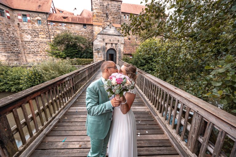 Zu sehen ist ein Brautpaar auf einer Hochzeit in Bayern, aufgenommen in Farbe, sie stehen auf einer Brücke und küssen sich. Sie halten den Blumenstrauß in die Kamera, im Hintergrund ist ein Schloss zu sehen