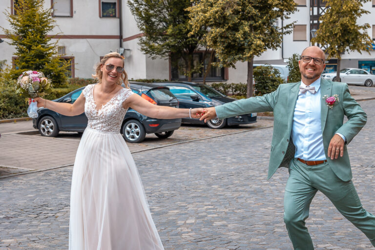 Ein Brautpaar tanzt ausgelassen nach der Trauung auf einer Straße in Bayern, aufgenommen in Farbe. Beide lächeln breit in die Kamera